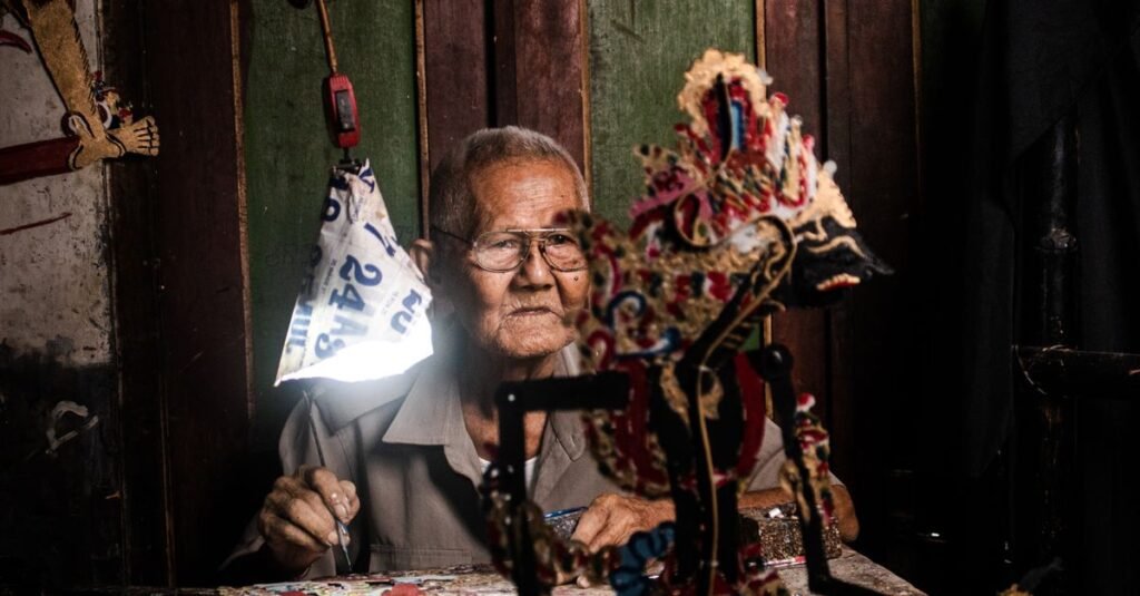 An elderly man crafts intricate wayang puppets in a traditional Indonesian setting.